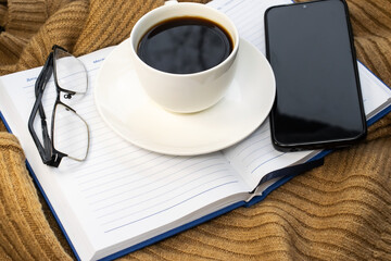 there is a white Cup of coffee on an open Notepad next to a mobile phone and eyeglasses.
