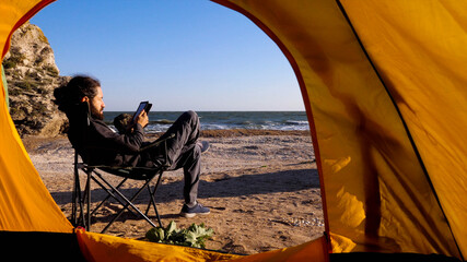 View from camping tent to bearded man sits on camp chair and reads electronic reader on the beach
