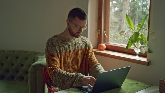 a man in a home sweater in a cozy home works on a laptop doing work remotely during the second wave of coronavirus