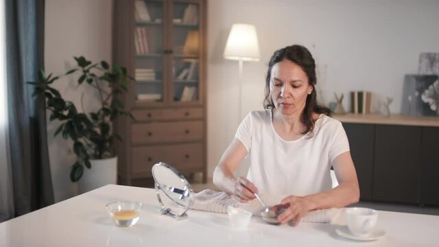 Medium Shot Of Attractive Middle Aged Woman Looking At Mirror While Applying Homemade Facial Scrub With Coffee Grounds Sitting In Cozy Living Room Taking Care Of Her Skin