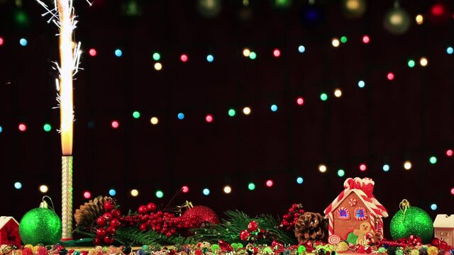 The fireworks are lit and throw white sparks upward. There are Christmas toys and a gingerbread house. In the background, colorful holiday lights flicker in the dark at night. New year greeting card.