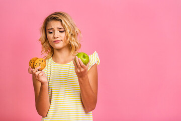 Diet concept. Portrait of cute little girl holding an apple and cookies isolated over pink background.