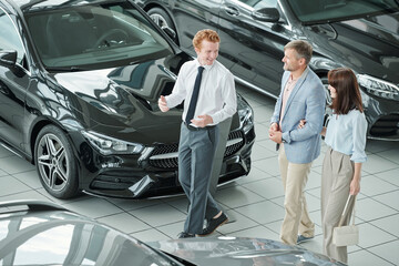 Confident young male manager of luxurious auto center consulting couple