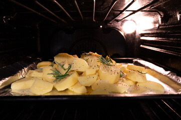 Close up shot of a pan inside the oven full of potatoes with salt, pepper and rosemary, the yellow light of the oven envelops them.