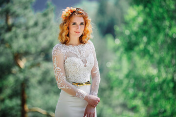 beautiful young red-haired bride in the forest with a floral wreath on her head. woman in long white dress outdoors on summer day. wedding day