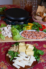 Raclette table setting with raclette machine and food ingredients. Swiss raclette served with different cheeses, fish, sald, mushrooms, paprika, bread. Self-service party.