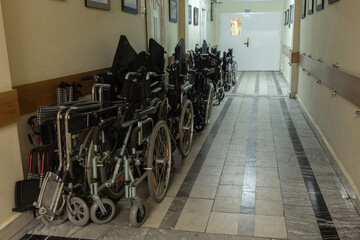 There are many wheelchairs on the worn marble floor in the corridor of an old hospital or nursing...