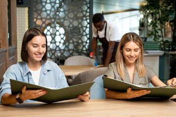 Two young smiling females sitting by table in cafe and looking through menu