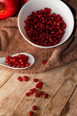 Overhead view of appetizing red pomegranate fruit kernels in a white porcelain bowl and spoon on top of a wooden table with a brown cloth and fruit kernels scattered across the table. 