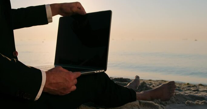 A man sitting on the beach in a suit, opens a laptop to start working, in the early morning against the background of the sea. Work while on vacation at sea.