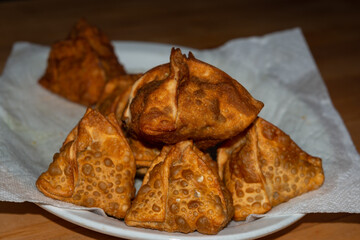 A plate of dark triangular fried samosa or baked pastry with a savory filling, such as spiced potatoes, onions, peas, cheese, beef and other meats, or lentils. The stack is on a white plate with paper
