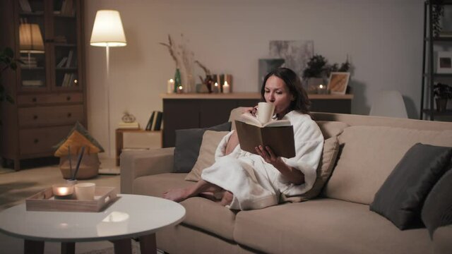 Medium shot of middle-aged woman in white bathrobe and towel reading book and drinking hot tea at night after doing beauty procedures smiling and enjoying time
