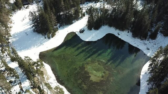 A drone shot of anAlpine valley in Austria. The valley has a Green Lake in the middle.  A headland in the middle of the lake. The algae give the lake distinctive color. Early spring in the mountains