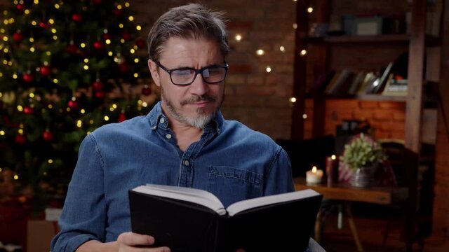 Man reading book at home at Christmas in winter. Happy older white man in glasses, smiling while reading, dark room with Christmas tree.