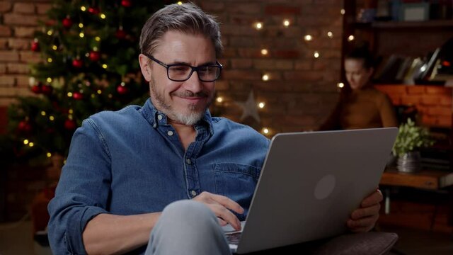 Older man working from home at Christmas in winter. Happy older white man with laptop at home, thinking, dark room with Christmas tree.