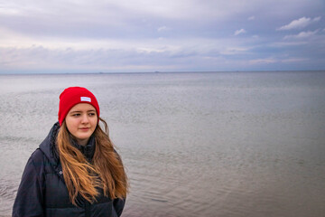a young girl with long hair, in a red cap, standing on the seashore, in a cool autumn morning
