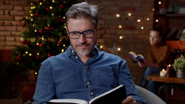 Couple reading books at home at Christmas in winter. Happy older white man in glasses, smiling while reading, dark room with Christmas tree.