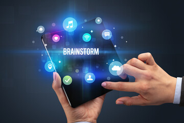 Businessman holding a foldable smartphone with BRAINSTORM inscription, social media concept