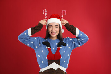 Fototapeta na wymiar Young woman in Christmas sweater and Santa hat holding candy canes on red background