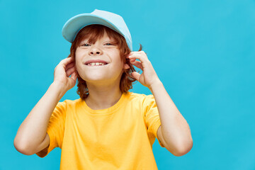 Cheerful red-haired boy holds his hands near the face blue cap yellow t-shirt close-up 