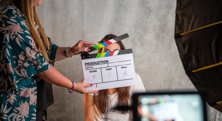 Operator holding clapperboard during the production of short film inside a studio with young actress on stage. Focus on the clapperboard and blur effect on the monitor and model. - obrazy, fototapety, plakaty