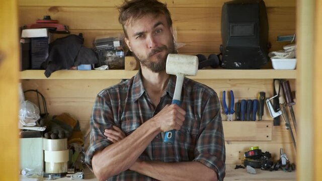 Funny brutal worker man stands in workshop and scratches his beard with a mallet, construction carpentry tools in the background. Portrait of cheerful craftsman. DIY concept.