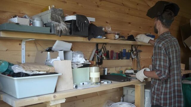 Tired craftsman returning to workshop, removes welding mask and gloves and choosing construction tools for next work.
