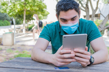 Obraz premium Young man with a protective mask watching multimedia content on a tablet, sitting in a park.