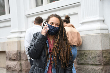 Female with long dread locks talks on phone while wear a face mask outside. 