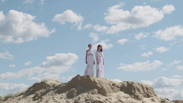 Two young girls in white Boho dresses pose together on a sand hill against a blue sky. Happy young people, teenage friendship