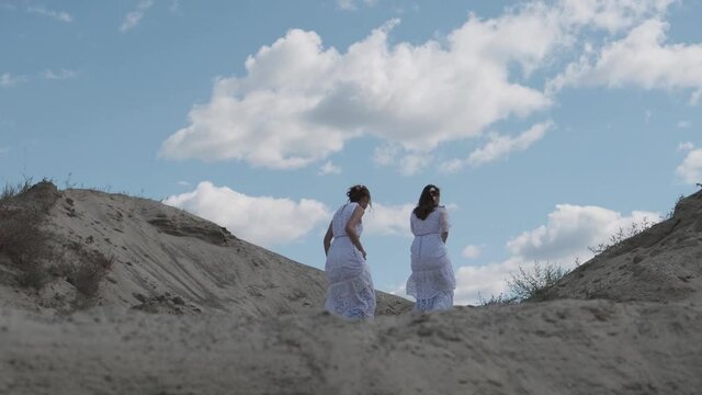 Two young girls in white boho dresses walk together on a sandy hill against a blue sky. Happy young people, teen friendships