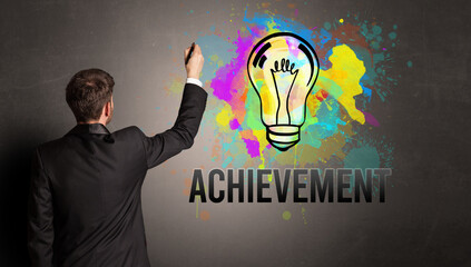 businessman drawing colorful light bulb with ACHIEVEMENT inscription on textured concrete wall, new business idea concept