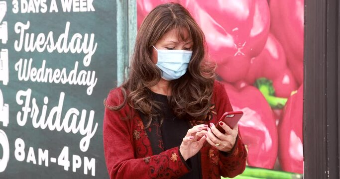 Attractive exotic-looking asian older woman with long curly hair wearing covid mask for coronavirus texts on cell phone leaning on rail.  Bright, hand-held medium shot with bokeh background.