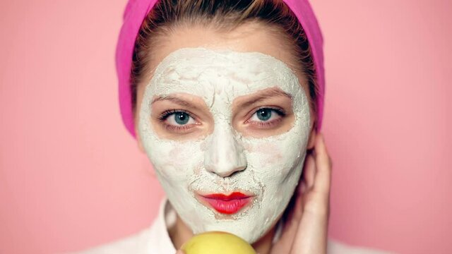 Close up of a girl with a face mask eats an apple on a pink background. Funny beautiful girl with cosmetic mask on her face. Woman wearing face mask. Face care concept.