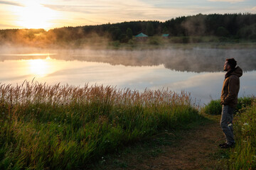 Fototapeta na wymiar A full-length man stands on the green shore of the lake. Beautiful sunrise, fog over the water.