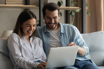 Happy millennial man and woman sit on sofa relaxing watch funny video on laptop laughing together. Smiling young Caucasian couple have fun rest look at computer screen enjoy online shopping from home.