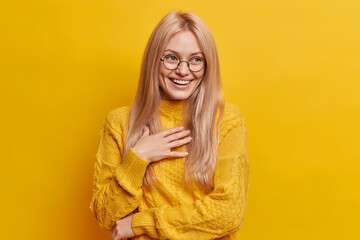 Joyful blonde European woman laughs happily looks carefree giggles positively keeps hand on chest...