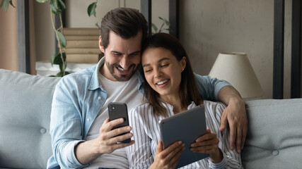 Wide banner panoramic view of happy millennial spouses man and woman relax on sofa using modern gadgets together. Smiling Caucasian couple rest at home have fun browsing electronic devices.