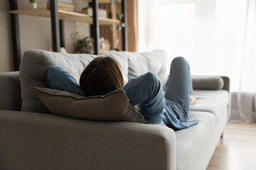 Back view of tired woman lying relaxing on comfortable sofa in living room take nap or daydream....