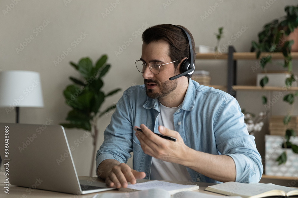 Wall mural Young Caucasian man in earphones sit at desk in home office have online video training or meeting on computer. Millennial male in headphones work distant talk speak on webcam digital call on laptop.