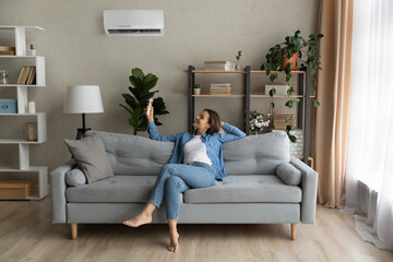 Happy young Caucasian woman relax on couch in living room turn on air conditioner with remote...
