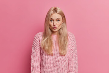 Fototapeta premium Lovely romantic woman with blonde hair keeps lips rounded wants to kiss boyfriend flirts with someone wears warm knitted sweater isolated over pink background. Human face expressions concept
