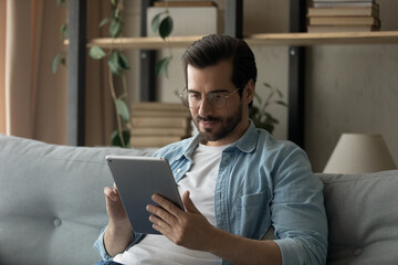 Young Caucasian man in glasses sit rest on sofa in living room look at tablet screen browsing wireless internet. Millennial male relax on couch at home shopping online on pad. Technology concept.