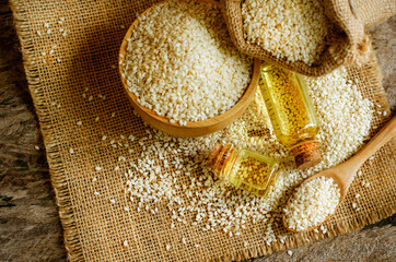 Dry white organic sesame seeds in wooden spoon with extract oil in glass bottle on sack fabric and grunge background in dark tone