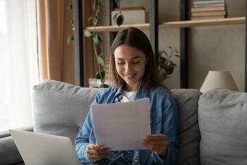 Smiling young Caucasian woman sit on sofa working on laptop read paper documents or correspondence. Happy female use computer at home get good pleasant news or notification in paperwork.