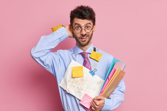 Puzzled office worker does paper work in home office scratches head with embarrassement prepares financial report wears formal clothes stands indoor. Male student hears exam or test results.