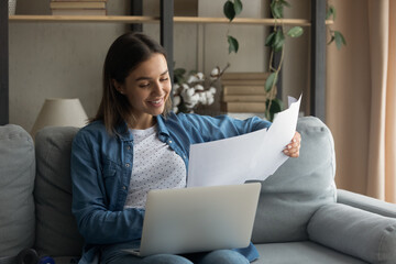 Happy millennial Caucasian female sit on couch study distant on laptop at home, read paperwork...