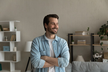 Fototapeta premium Happy young Caucasian man stand look in window distance thinking or planning future success. Smiling millennial male feel excited at home dreaming or visualizing opportunities. Vision concept.