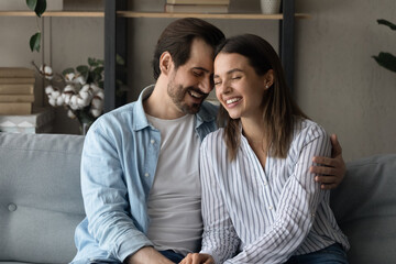 Naklejka premium Happy millennial man and woman married spouses sit on sofa at home hugging cuddling. Smiling young Caucasian couple embrace enjoy tender intimate romantic moment together. Love, relations concept.