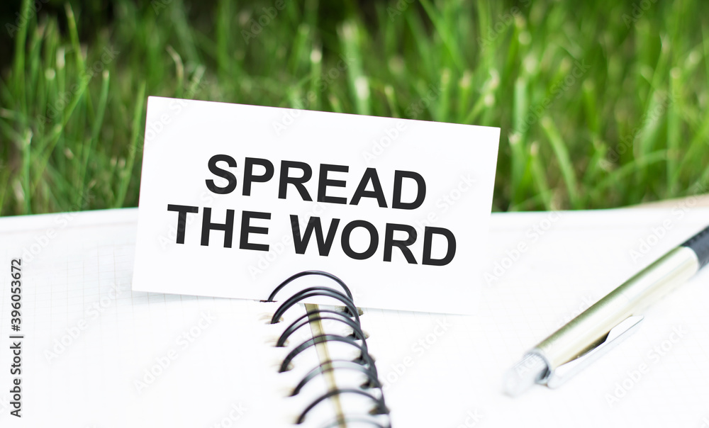 Poster text spread the word on a white card that lies on an open notebook on the table, next to the pen.